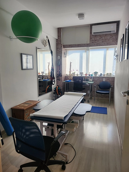 imagem ampla do consultório, com a mesa de atendimento, ao centro, bola de pilates e outros instrumentos